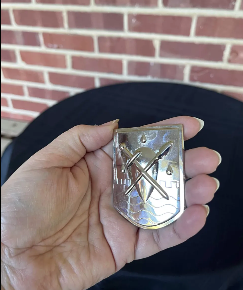 Silver plated Liscor City Watch badge held in the palm: a shield with a crossed sword and spear over raised waves, castellations and three raindrops