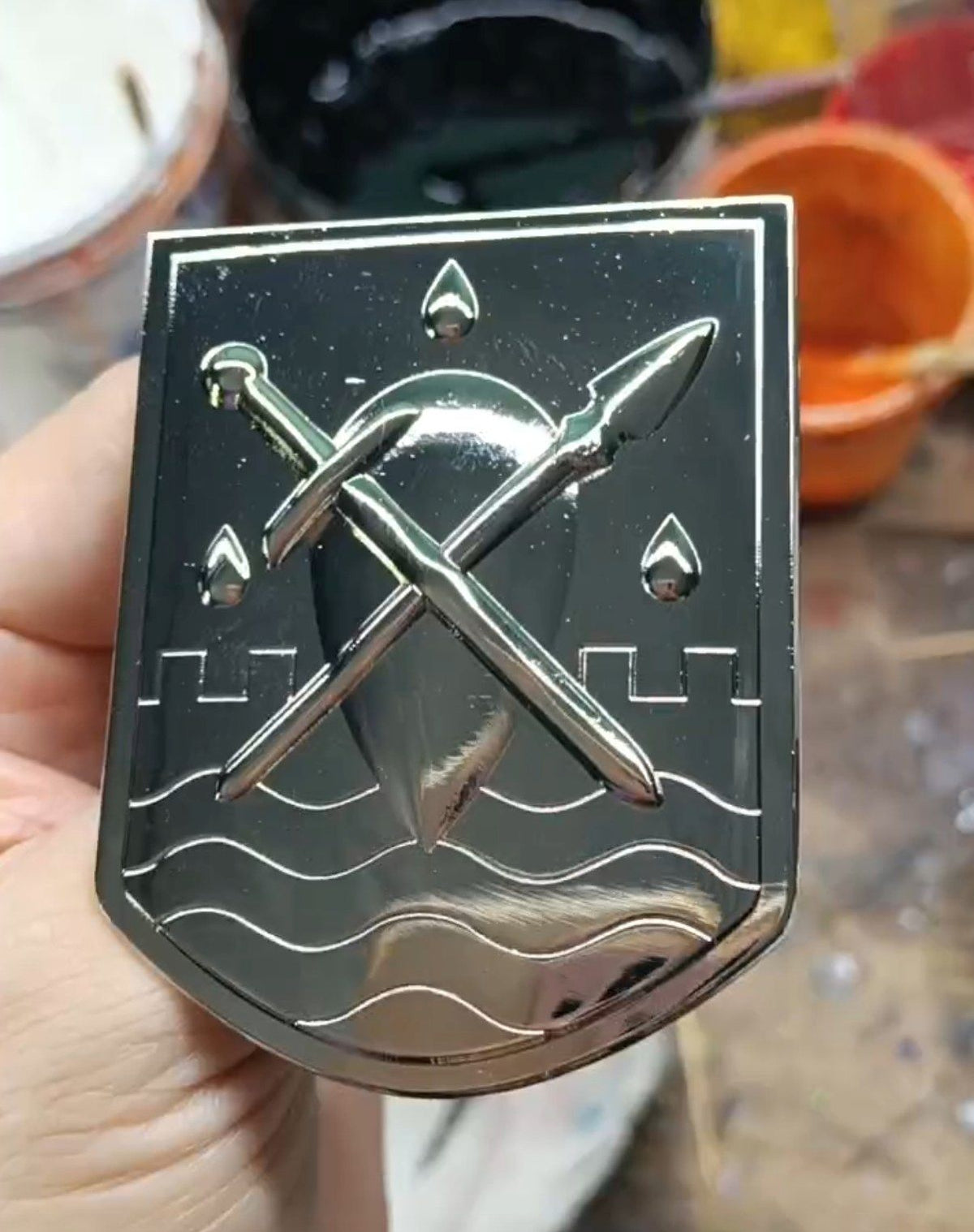 Silver plated Liscor City Watch badge held in one hand: a shield bearing a crossed sword and spear over raised waves, with castellations and three teardrops