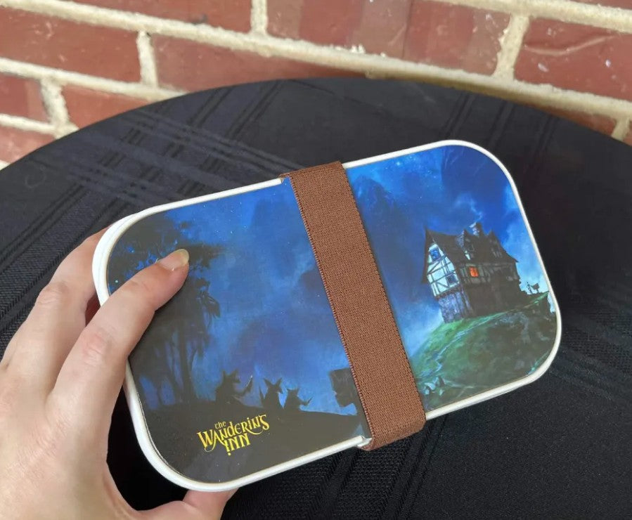 A hand holding The Wandering Inn bento lunch box, illustrated lid with elastic strap