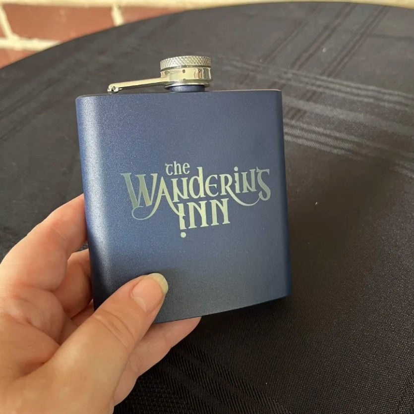 A hand holding The Wandering Inn stainless steel hip flask, showing the logo