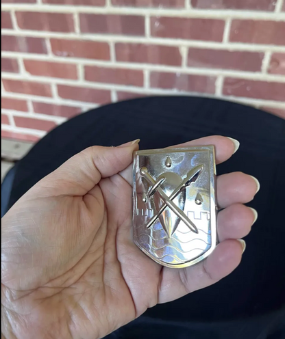 Silver plated Liscor City Watch badge held in the palm: a shield with a crossed sword and spear over raised waves, castellations and three raindrops