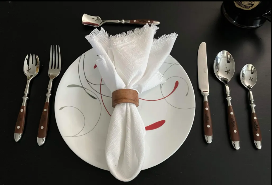 Six-piece Wandering Inn cutlery set laid out around a plate: dinner fork, dinner knife, large spoon, small spoon, spork and butter spreader, each with a dark wooden handle and stainless steel head