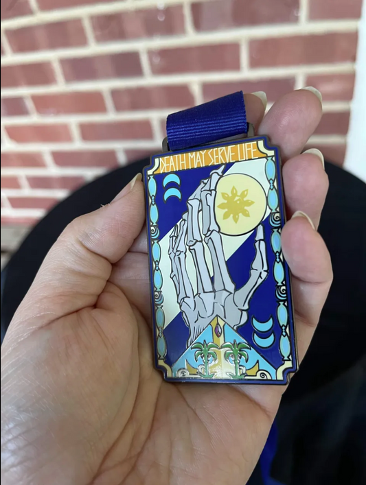 Khelt medallion held in hand: a skeletal hand reaching for a yellow sun in blue and turquoise enamel, under the words Death May Serve Life