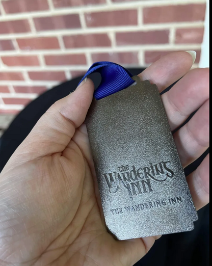 Reverse of the Khelt medallion: The Wandering Inn logo and wordmark raised on the textured metal back