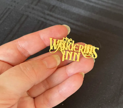 The Wandering Inn logo lapel pin held in hand, the gold-plated wordmark cut as a single piece