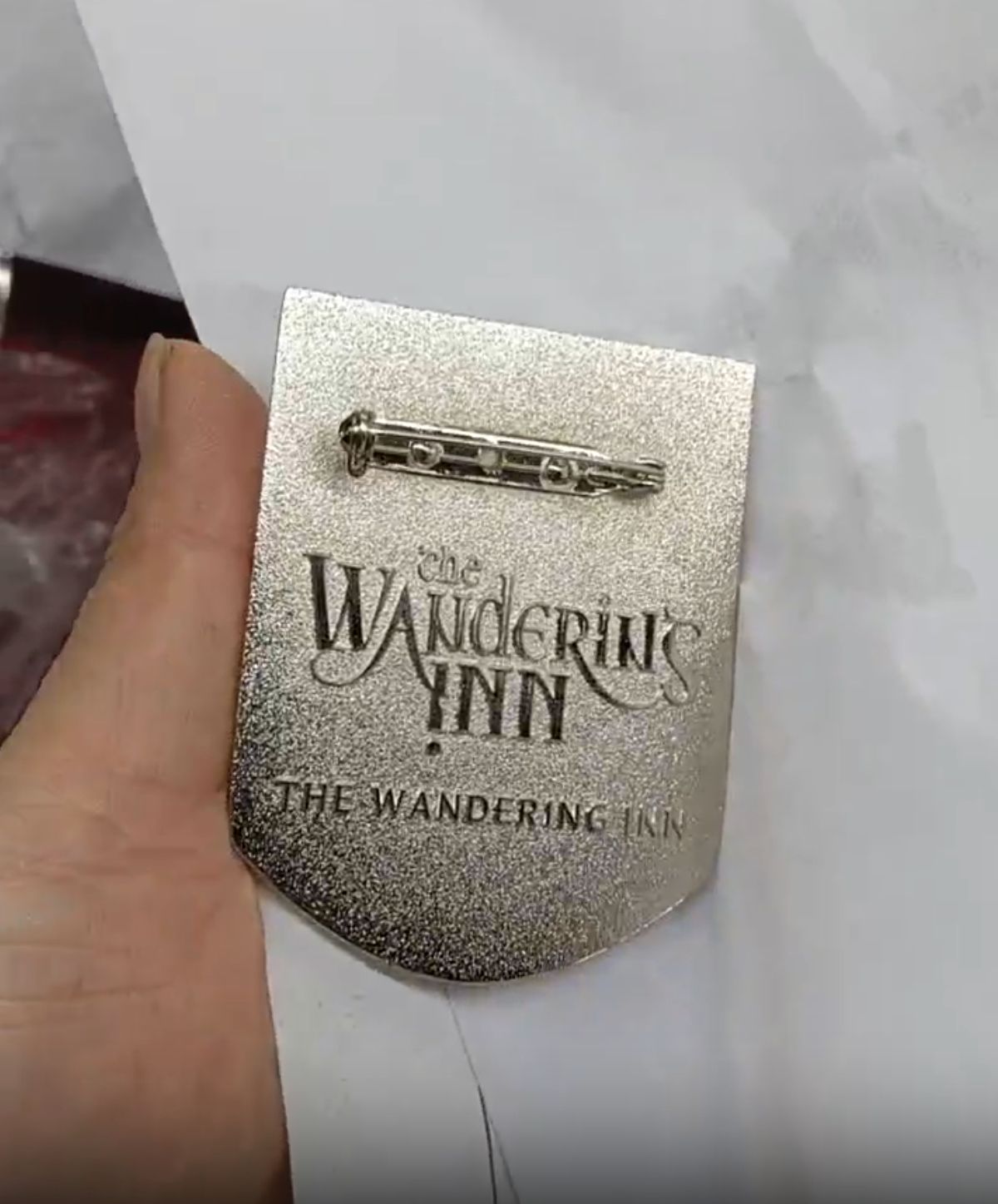 Reverse of the Liscor City Watch badge showing the pin fastening and The Wandering Inn logo engraved into the brushed metal back